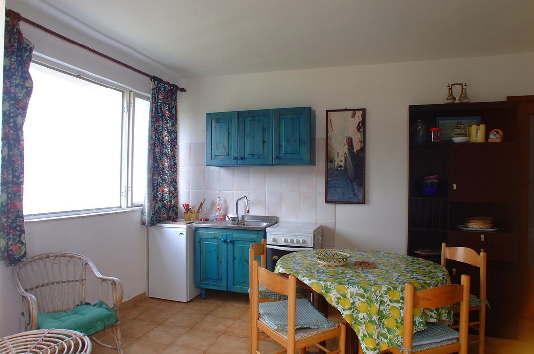 A compact kitchen-dining area with turquoise cabinetry, small appliances, wooden dining table, and large window with floral curtains.