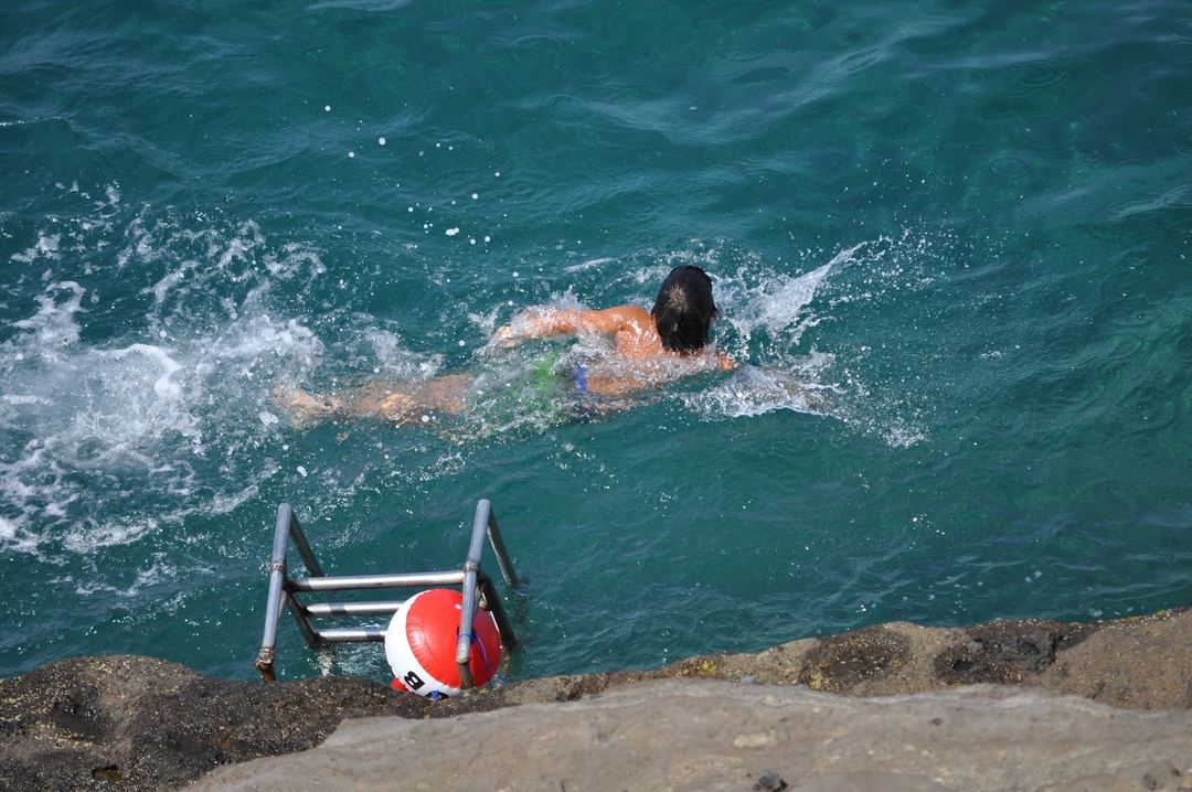 A swimmer performs freestyle strokes in turquoise Mediterranean waters near a rocky beach with a ladder and buoy in the foreground.