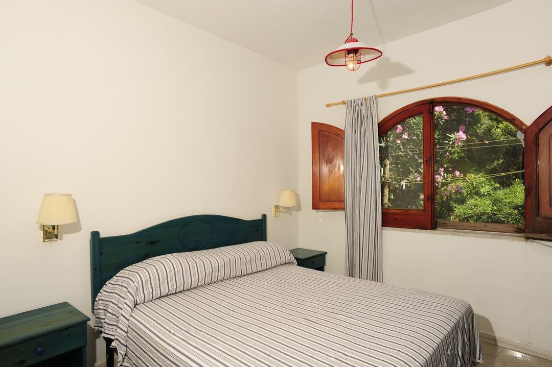 A bright bedroom with a teal-framed bed, striped bedding, wall sconces, and arched wooden doors with views of flowering gardens.