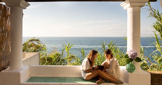 Two guests relax on a seaside terrace with classical columns overlooking the Mediterranean Sea.