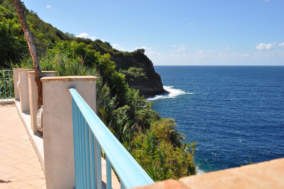 A scenic coastal viewpoint with a light blue railing overlooking dramatic cliffs covered in lush vegetation and sparkling blue ocean waters.